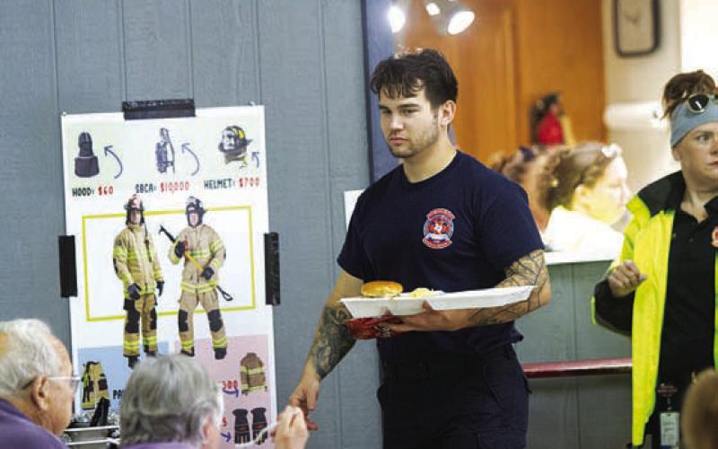 Caleb Doll walks by with a plate of food. At left, a sign can be seen showing the cost to outfit a firefighter. Funds raised from Saturday’s event will go toward equipment upgrades. HUNTER KING | DISPATCH RECORD